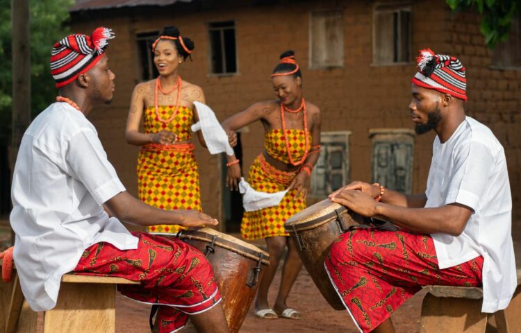 Men and women wearing Igbo traditional wear. The men are drumming while the ladies dance together happily smiling.