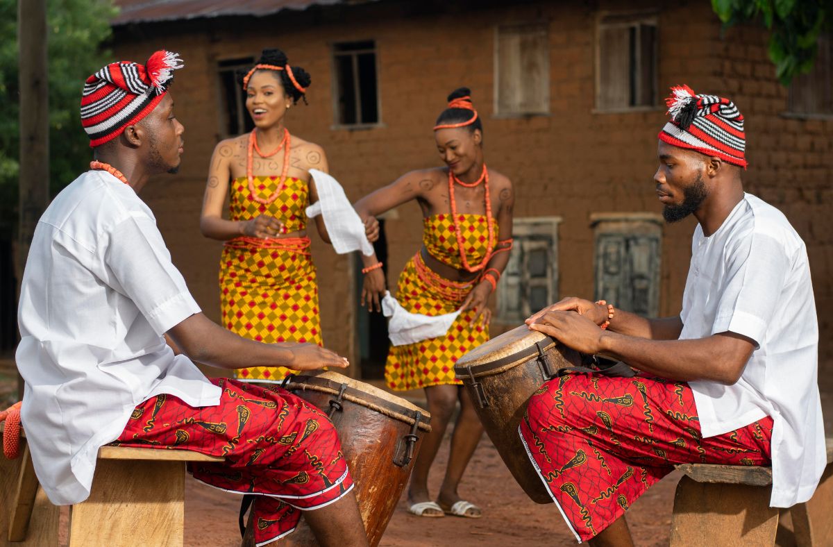 Men and women wearing Igbo traditional wear. The men are drumming while the ladies dance together happily smiling.